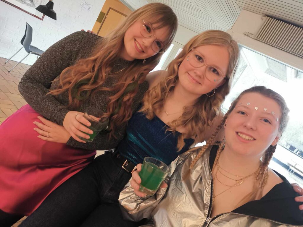 Tre smilende unge kvinder til festlig aktivitet med grønne drinks på en ungdomshøjskole.